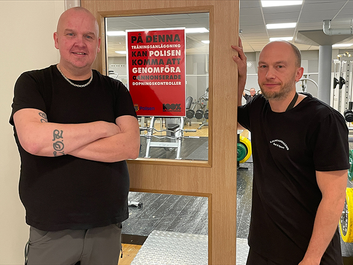 Två män i svarta tröjor, båda personal vid Ljungandalens bad och sport, står bredvid en skylt som visar att kommunen jobbar mot dopning.