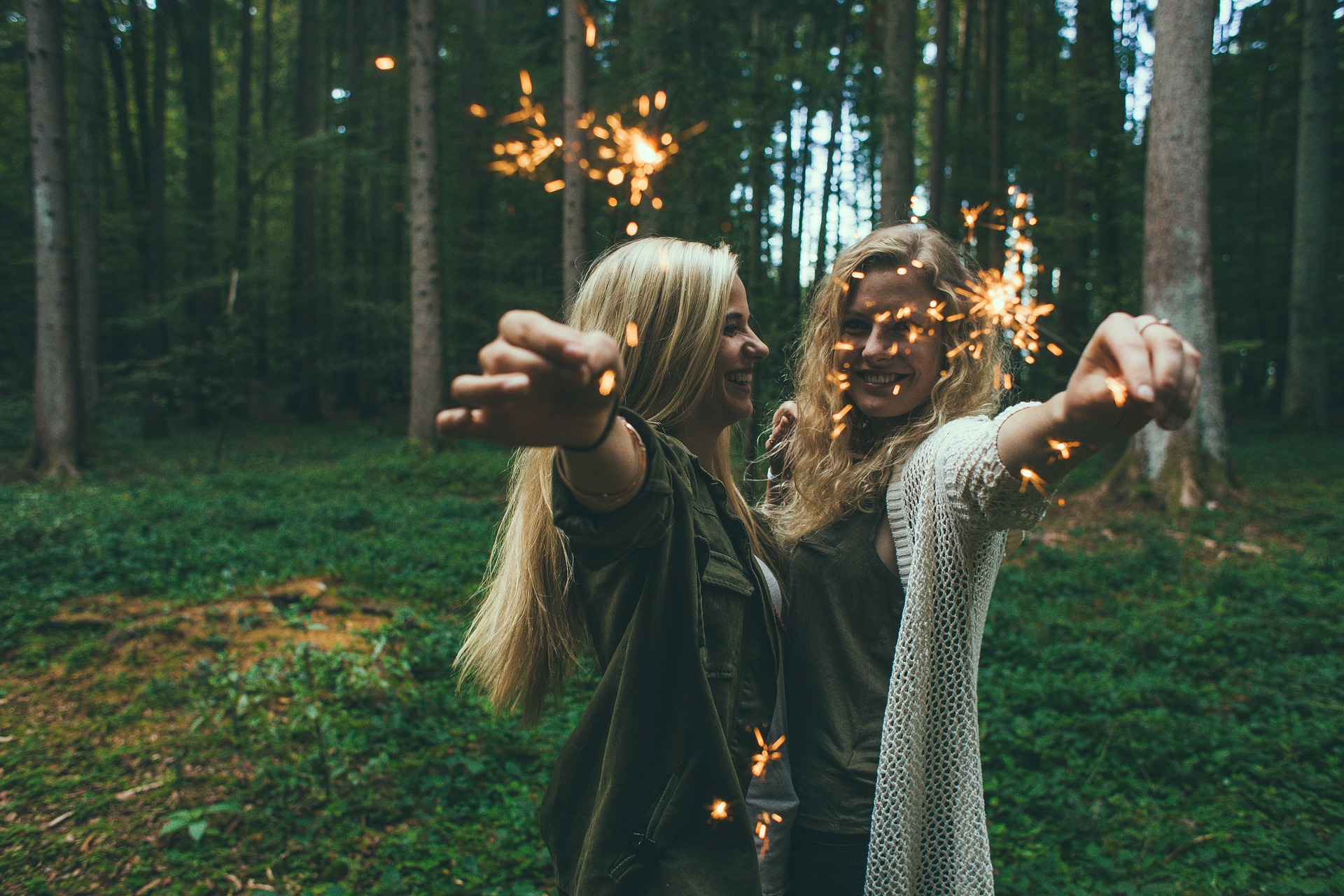 Bilden föreställer två glada tjejjer i en skog en midsommarnatt som håller i ett varsitt brinnande tomtebloss mot kameran.