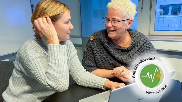 Två kvinnor sitter vid ett skrivbord med en dator framför sig. Kvinnan till vänster är ljushårig och har en vit tröja. Kvinnan till höger har grått hår och en mörk poncho. De tittar på varandra och ler.